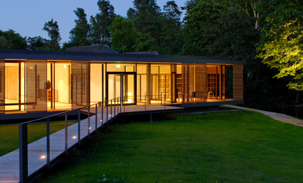 Blick auf innenbeleuchtetes Seminargebäude Gut Siggen mit Steg über Rasen und Bäumen ium Hintergrund bei Dämmerung