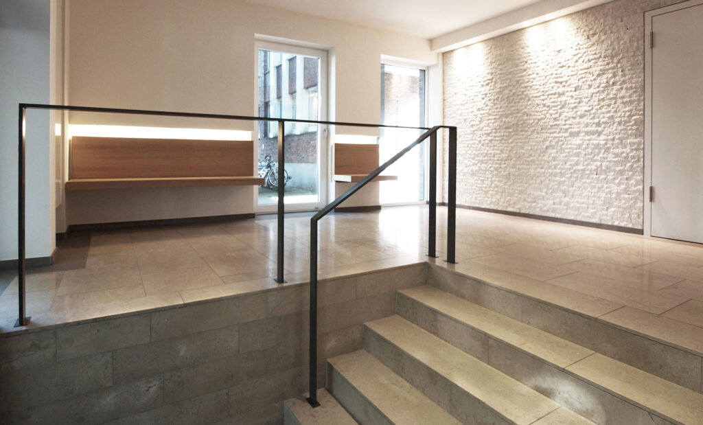 kurze Treppe mit Metallgeländer in einem weiß gehaltenem Foyer mit Holzbänken an der Seite
