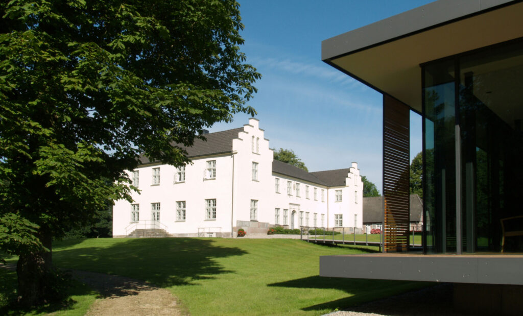 Außenansicht weißes Herrenhaus Gut Siggen, davor Wiese mit Bäumen und ein Nebengebäude im Anschnitt