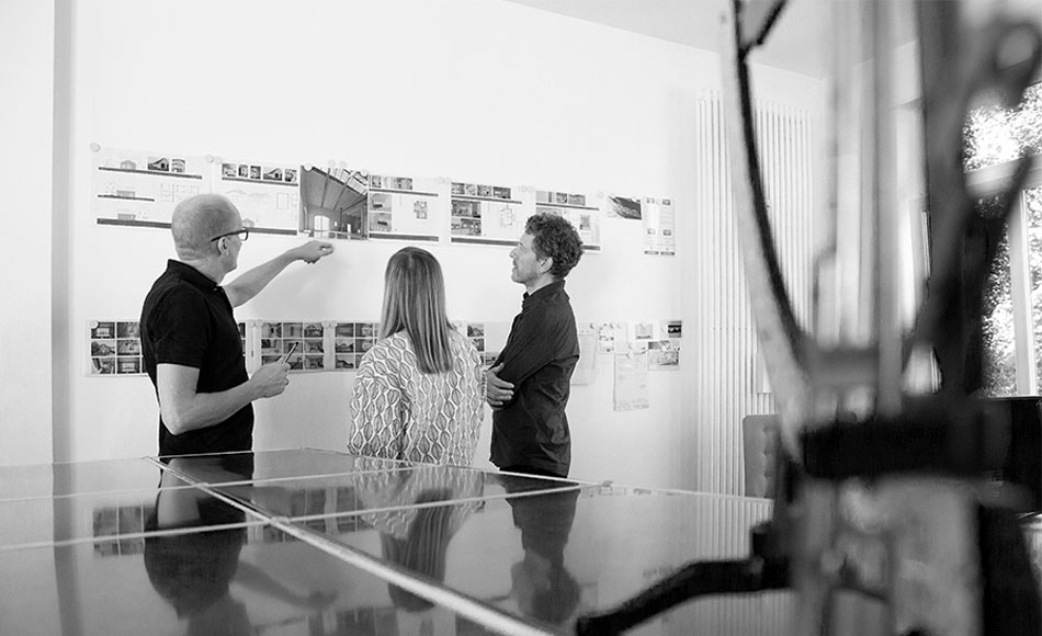 Drei Personen besprechen Architekturzeichnungen vor einer Wand.